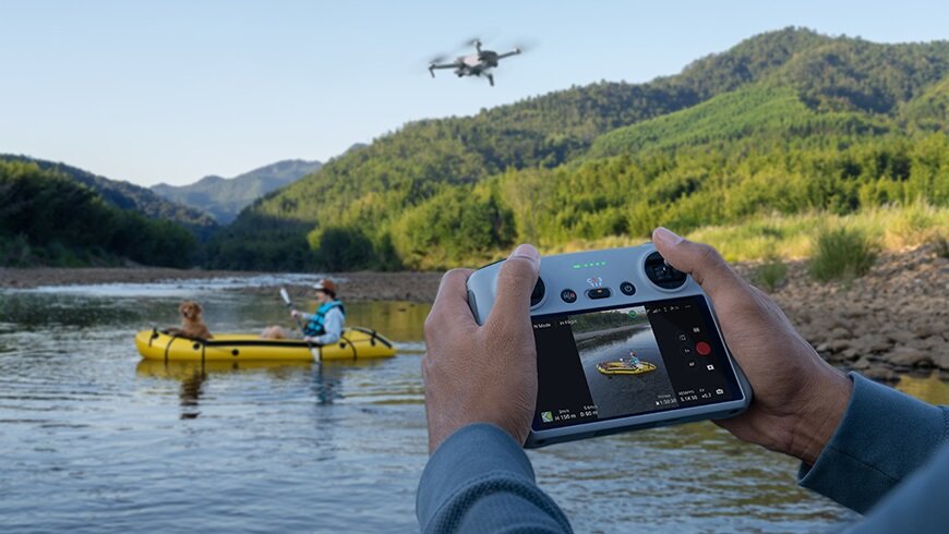 Dron DJI Mini 3 Fly More Combo CP.MA.00000613.01 widok na kontroler RC trzymany w dłoniach pod skosem w lewo oraz drona latającego nad wodą i mężczyznę z psem na pontonie