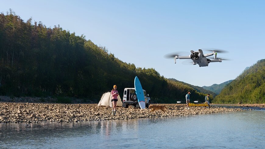 Dron DJI Mini 3 Fly More Combo CP.MA.00000613.01 latający nad wodą oraz sterująca nim kobieta stojąca na kamieniach przy brzegu